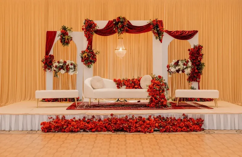 Grand red and gold reception stage with white arches, red velvet swags and red rose florals by Events By Maryam