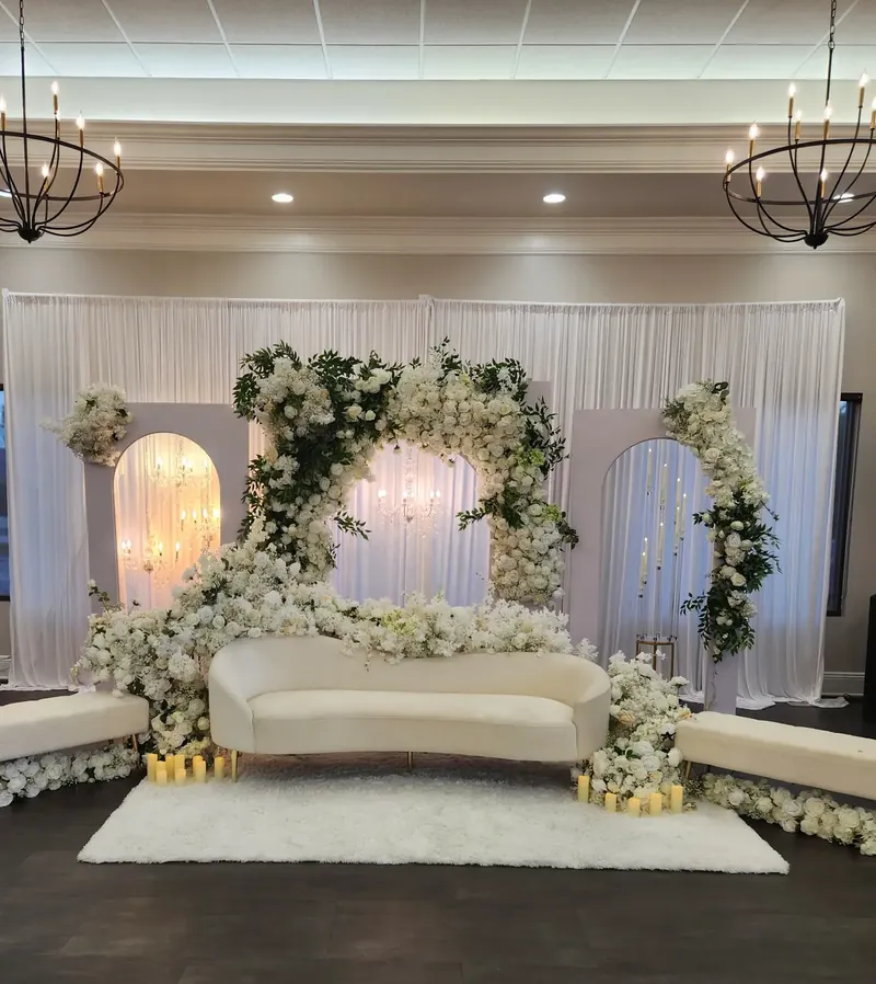 White floral arch stage with lit arch panels, a curved ivory sofa and iron chandeliers by Events By Maryam