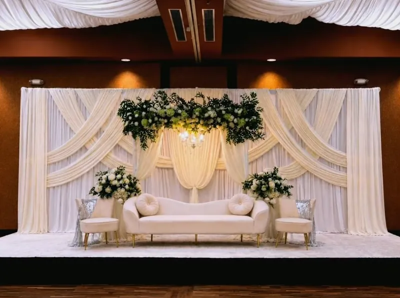 Ivory swag-draped grand stage with greenery canopy, chandelier and ivory sofa by Events By Maryam