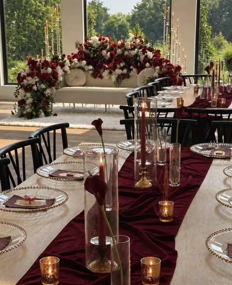 Burgundy velvet tablescape with calla lilies and gold-beaded chargers before a red-and-white floral sweetheart sofa by Events By Maryam