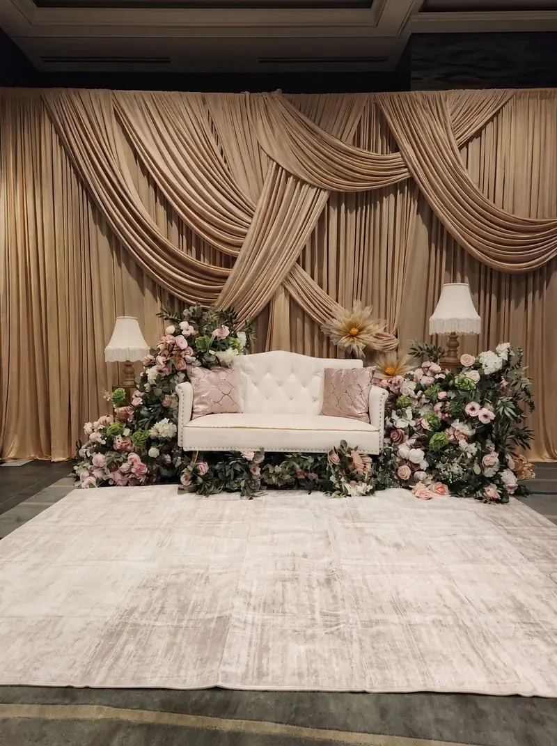 Champagne swag-draped backdrop with a tufted ivory sofa, blush florals and fringed lamps by Events By Maryam