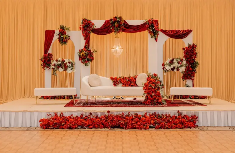 Grand red and gold wedding stage with white arches, red velvet swags, a crystal chandelier and red rose florals by Events By Maryam