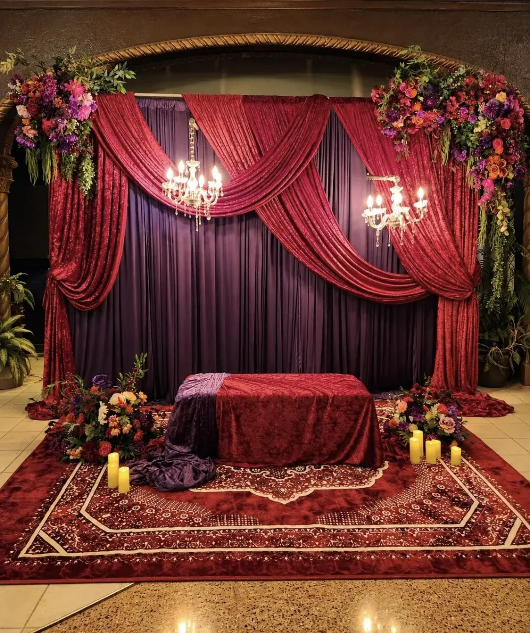 Burgundy and plum velvet draped backdrop with crystal chandeliers, jewel-tone florals and candles by Events By Maryam