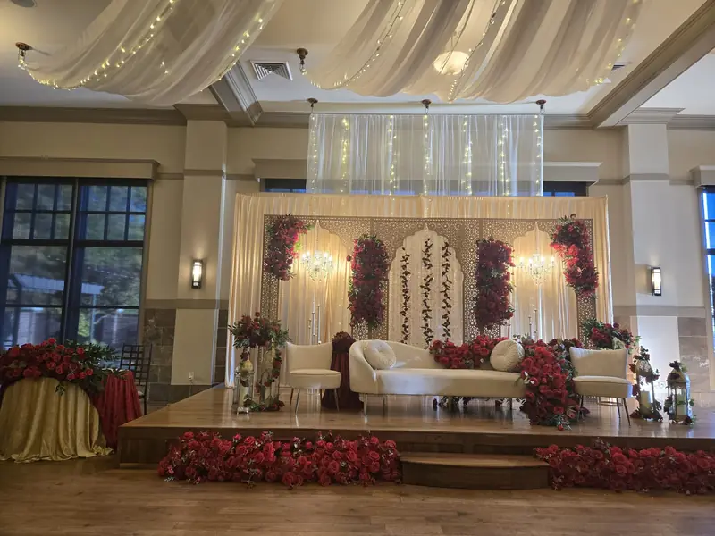 Grand wedding stage with red rose florals, carved ivory panels, crystal chandeliers and draped ceiling swags by Events By Maryam