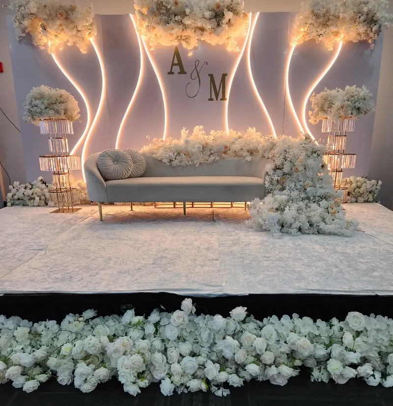 White floral stage with curved light panels, crystal stands, a curved sofa and a white rose border by Events By Maryam