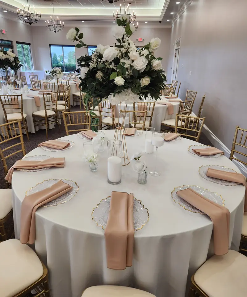 Guest table with a tall white rose centerpiece, blush napkins and gold chiavari chairs by Events By Maryam